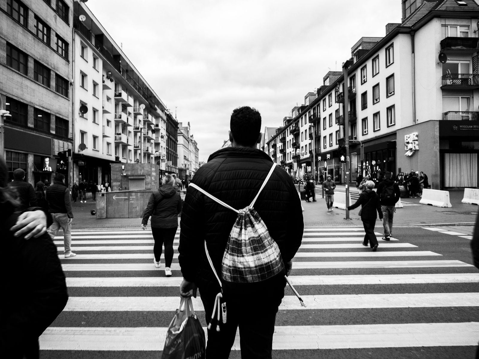 Black and white street photo of a person with a plaid backpack walking across a zebra crossing between tall buildings