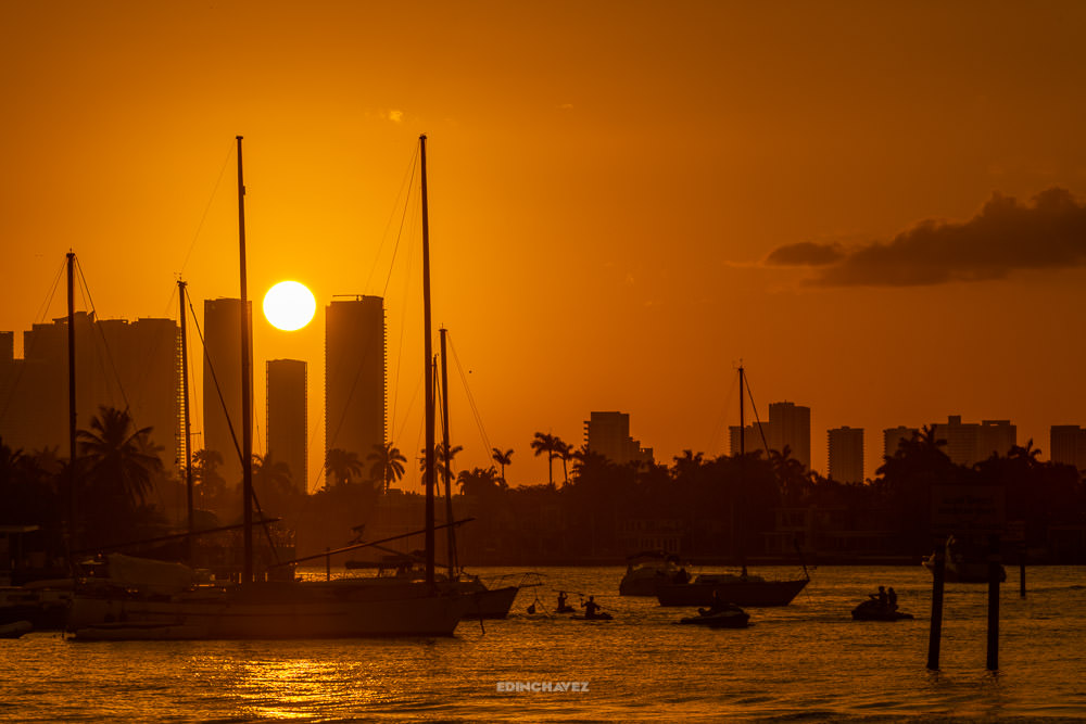 Sailboats and Miami buildings silhouetted against an orange sunset over the water