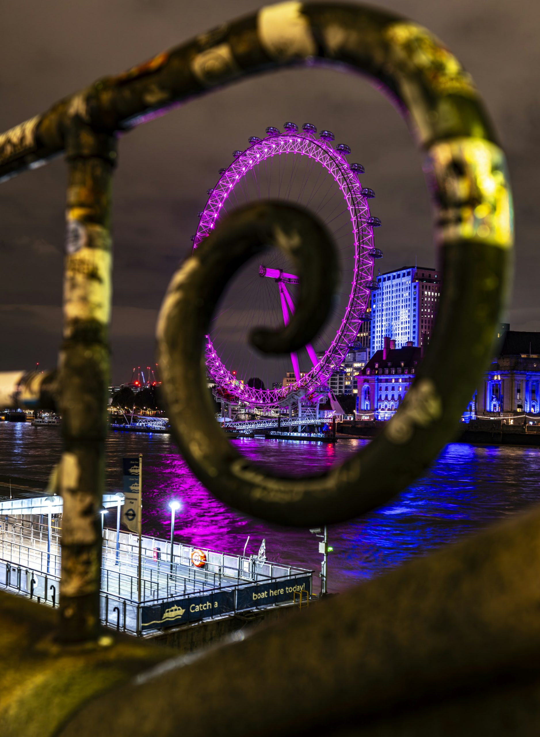 London Eye illuminated in purple above River Thames reflections, viewed through a blurred railing loop