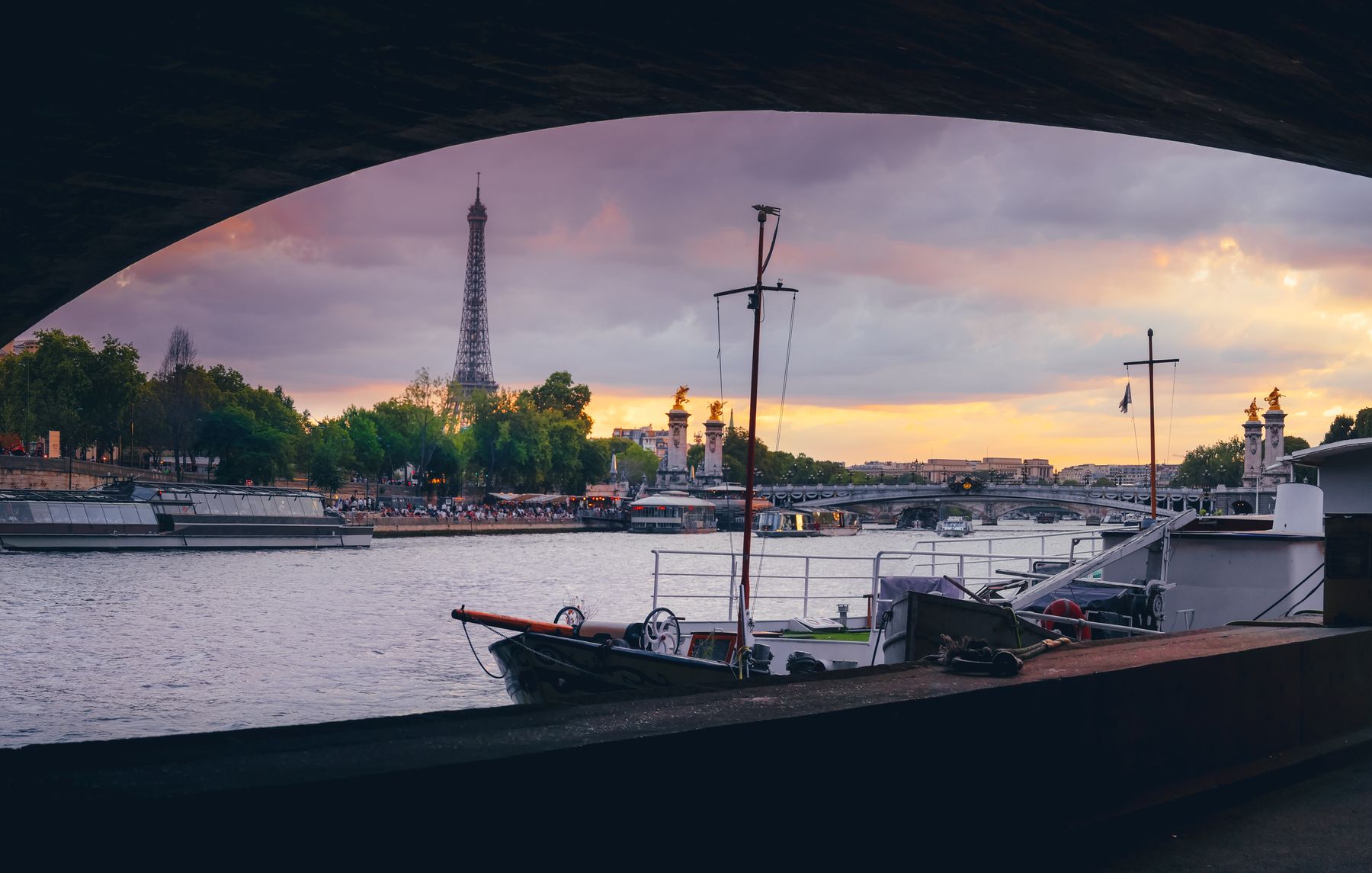 The Seine at dusk framed by the dark curve of a stone bridge arch, with moored boats and the Eiffel Tower in the distance