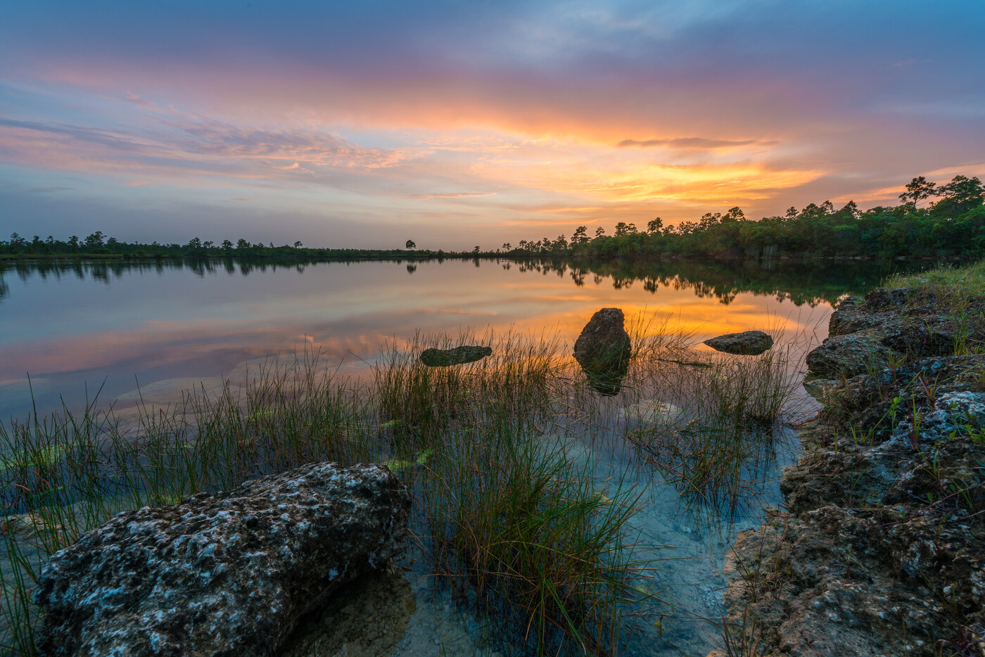 A calm lake at dusk with grass and rocks in the foreground and a blue, orange and purple sky reflected in the water