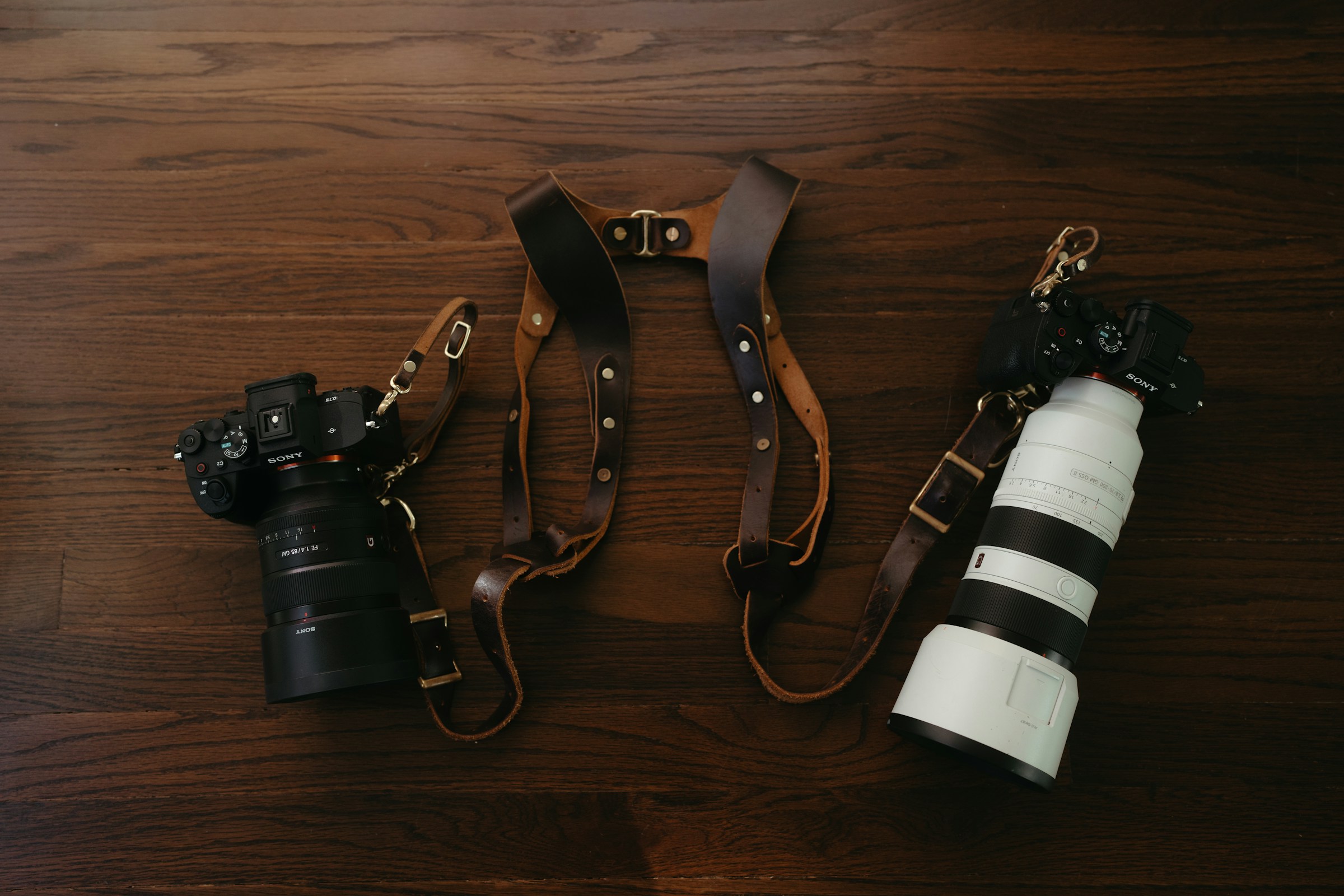 Two cameras with lenses resting on a brown leather dual camera harness on a wooden floor