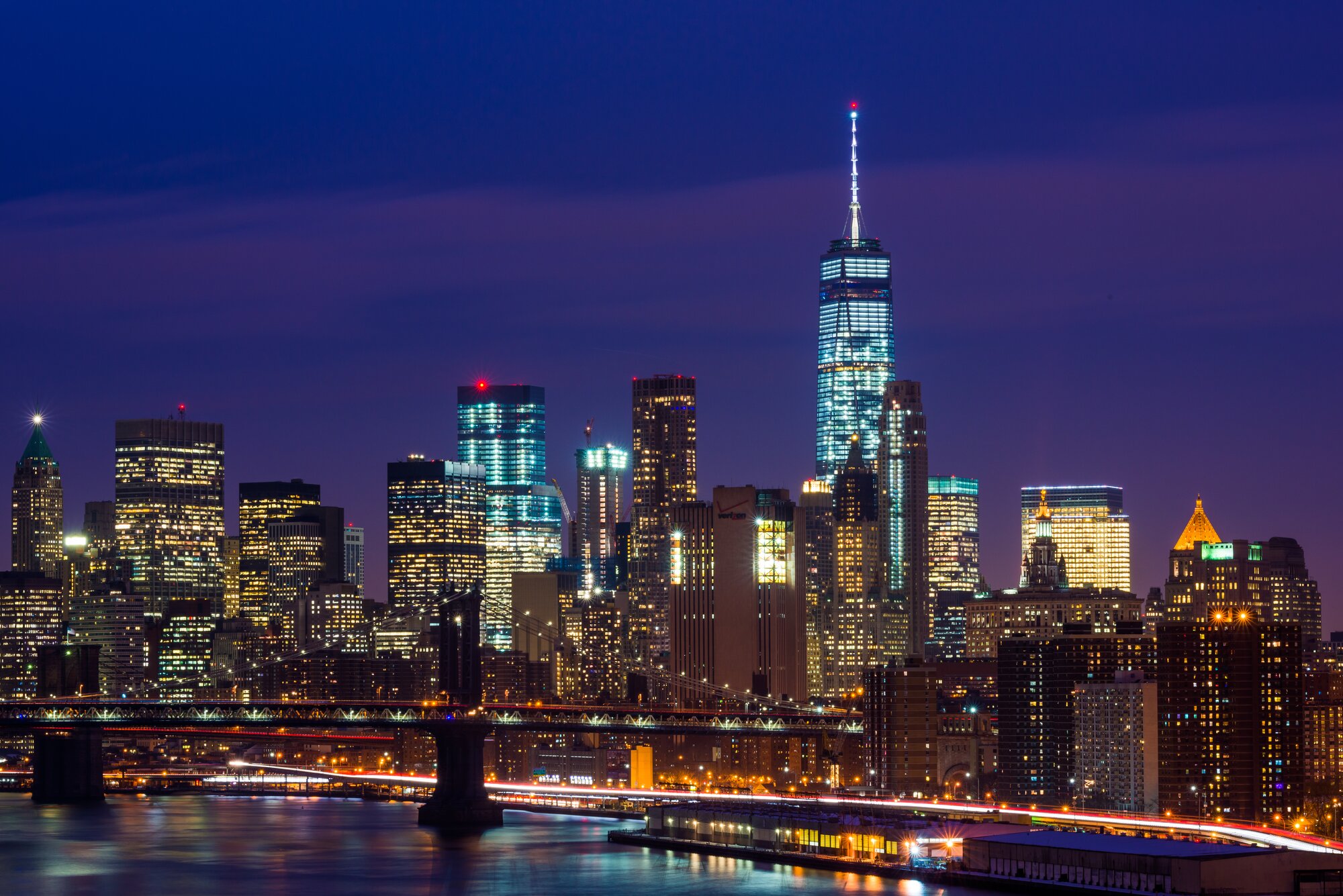 Cityscape photo of the lower Manhattan skyline and a bridge at blue hour with light trails