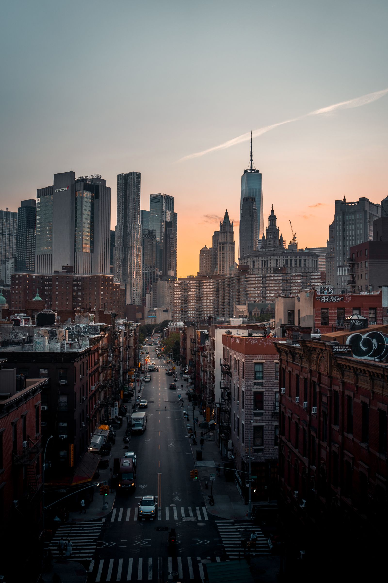 New York City street running toward the downtown skyline with One World Trade Center under a pink and orange dawn sky