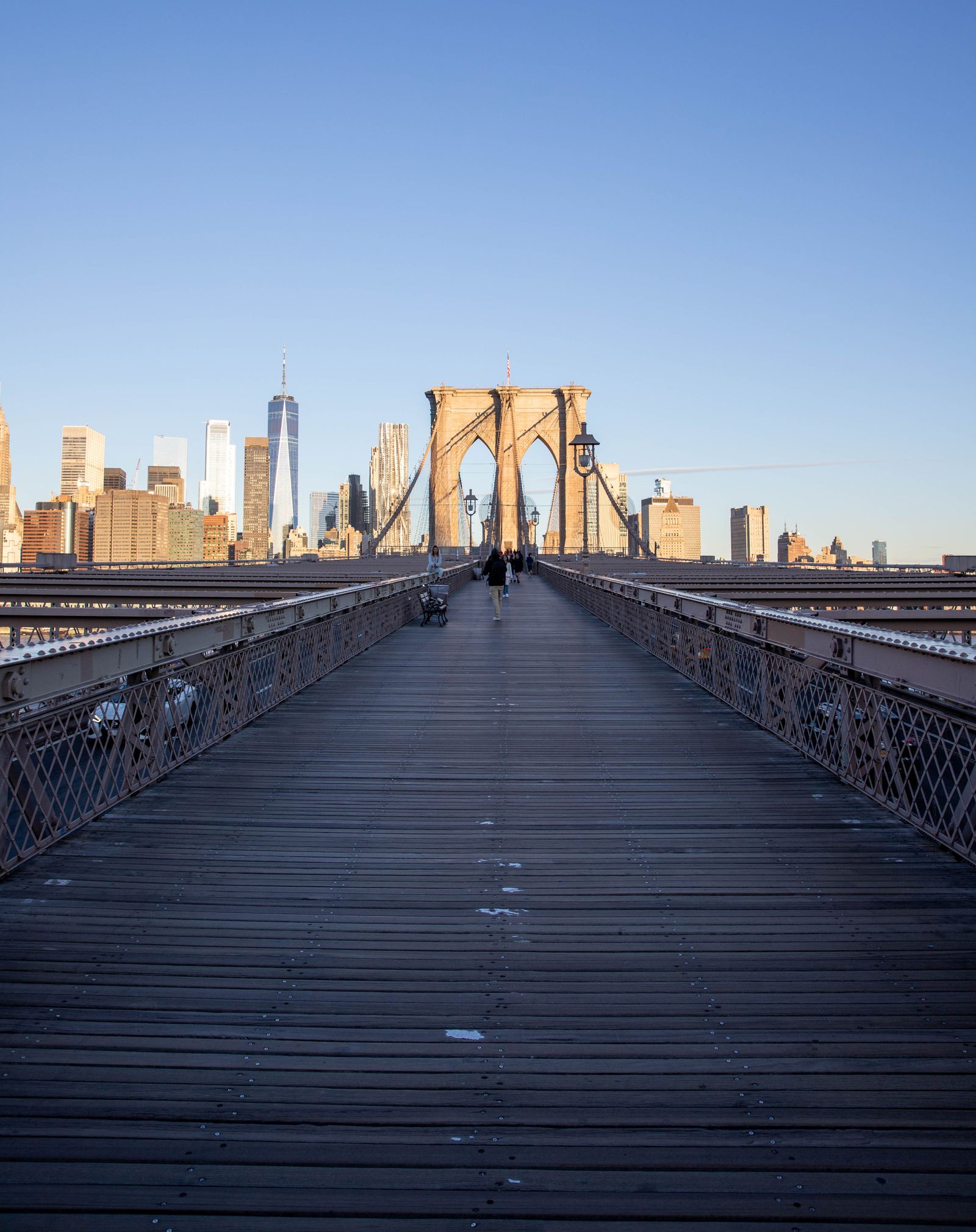 The Brooklyn Bridge walkway leading toward the Manhattan skyline with One World Trade Center under a clear morning sky