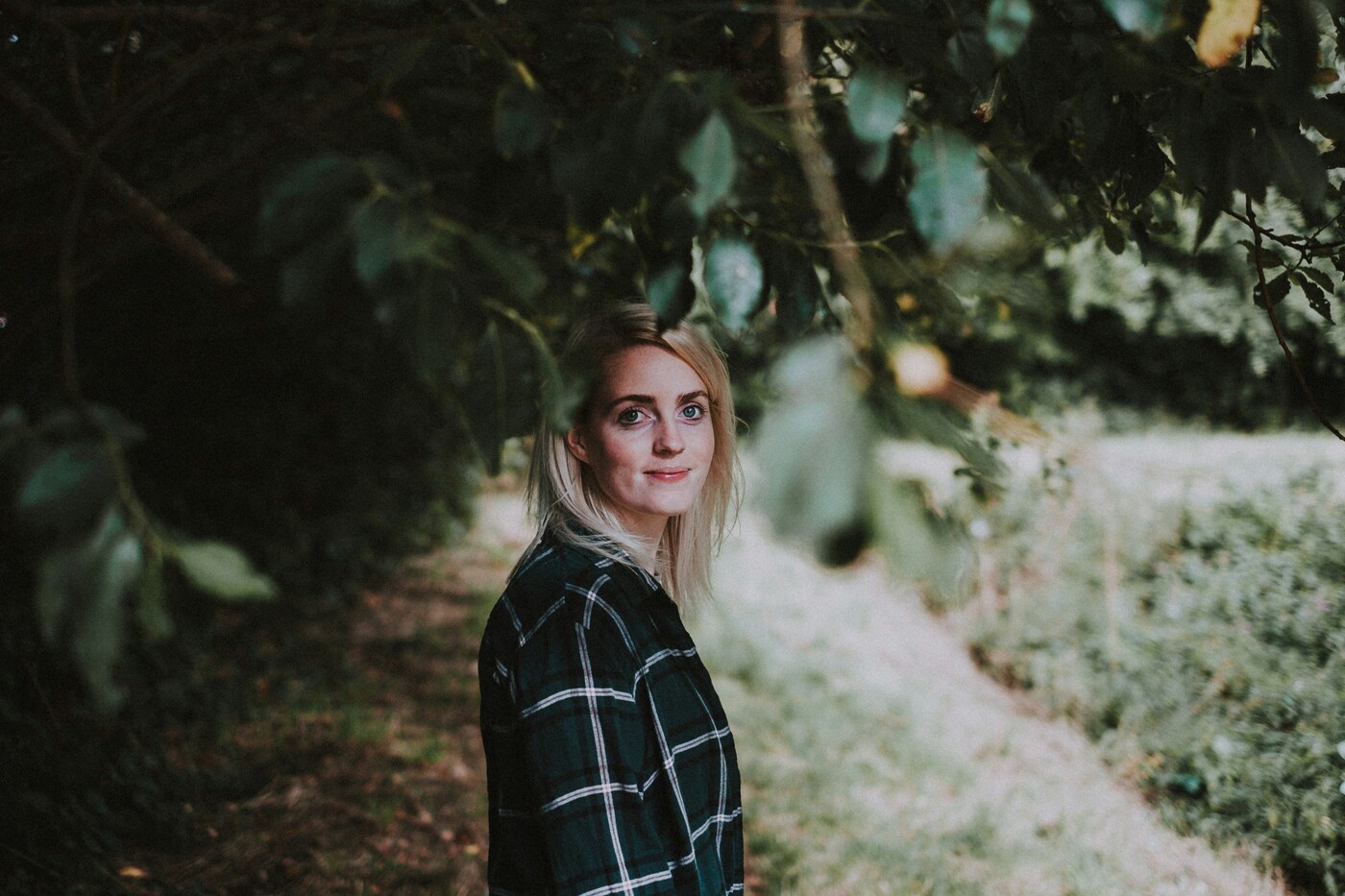 A woman with blond hair stands at the edge of a path under trees and looks at the camera in soft, shaded light