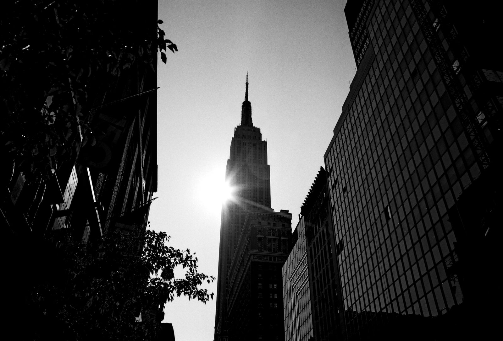 Black and white view of the Empire State Building silhouetted against a bright sun flare between dark buildings