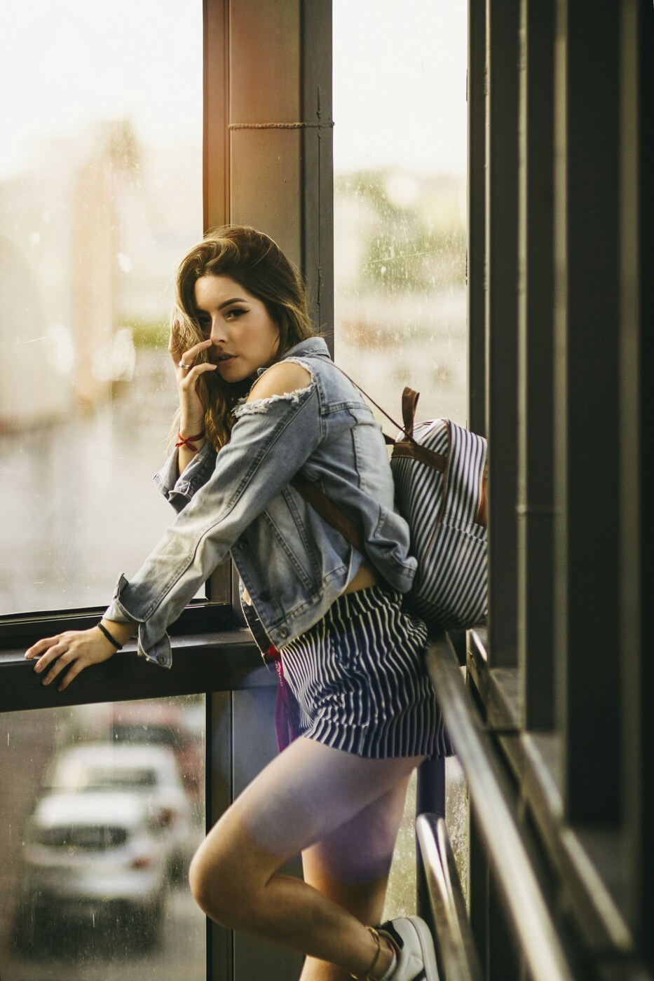 A woman leans on a window frame with her face turned toward the camera and a bright window behind her