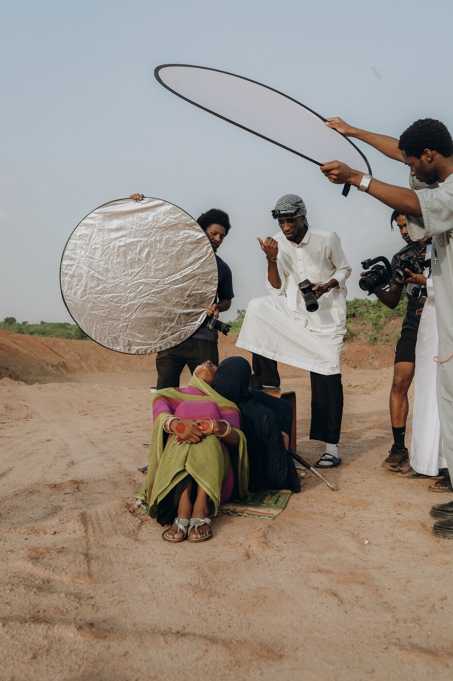 A crew outdoors on sandy ground with two round reflectors, one held above people seated on the ground