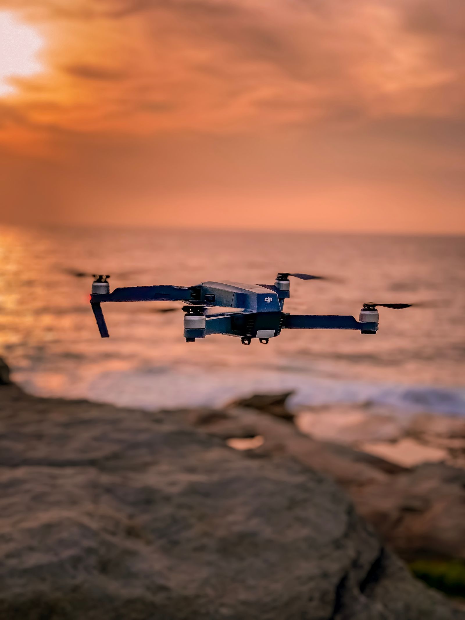 A small foldable camera drone hovering in the air over a rocky shoreline under an orange sunset sky