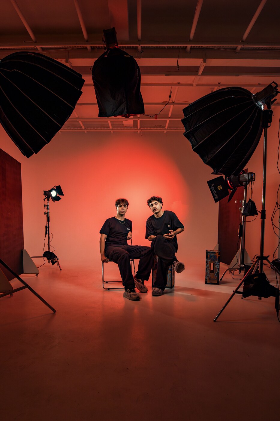 Two people sit on chairs in a studio lit in red, with softboxes and lights on stands around them