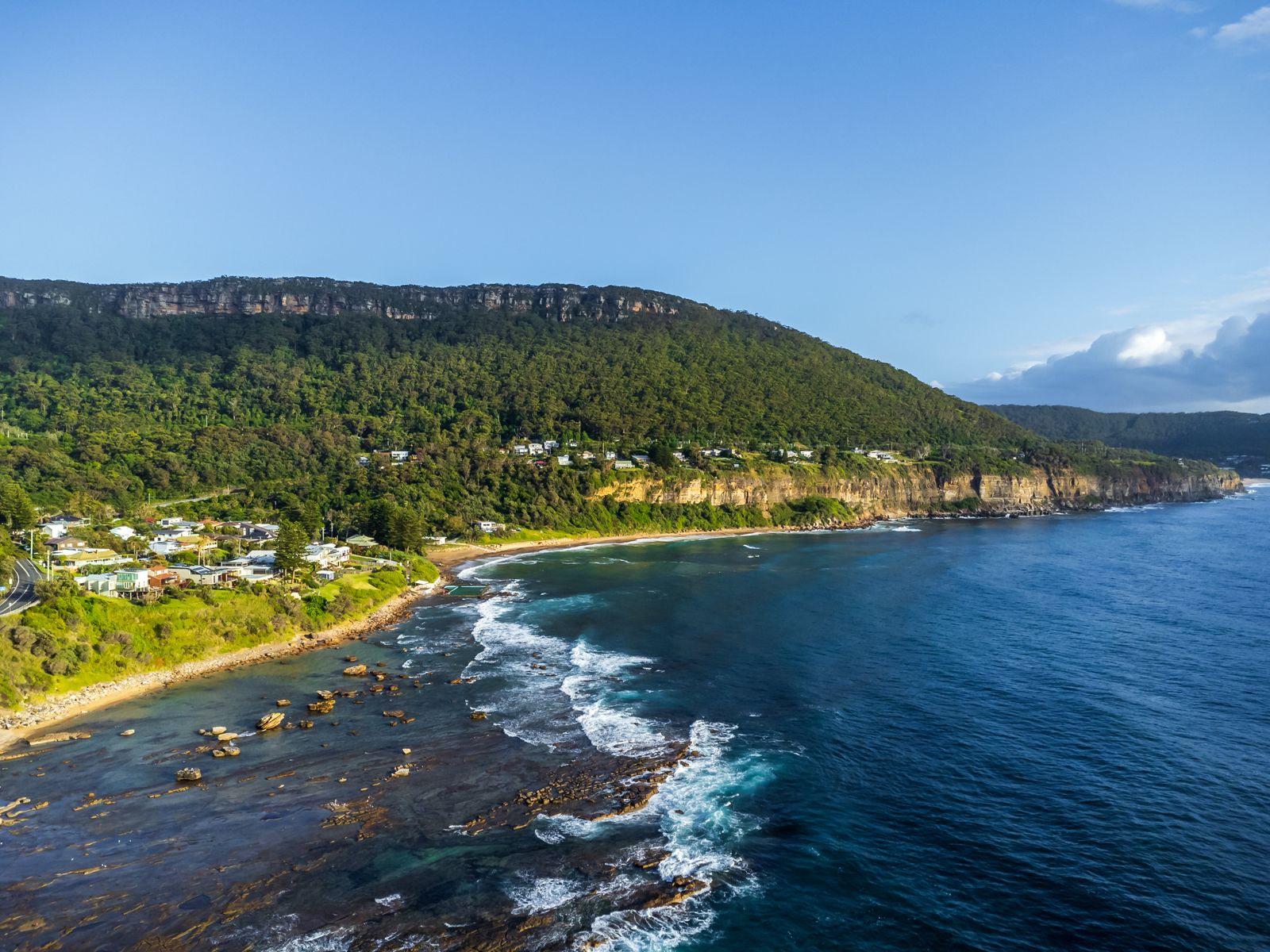Aerial view of a forested headland above a bright blue ocean with waves breaking on rocks below