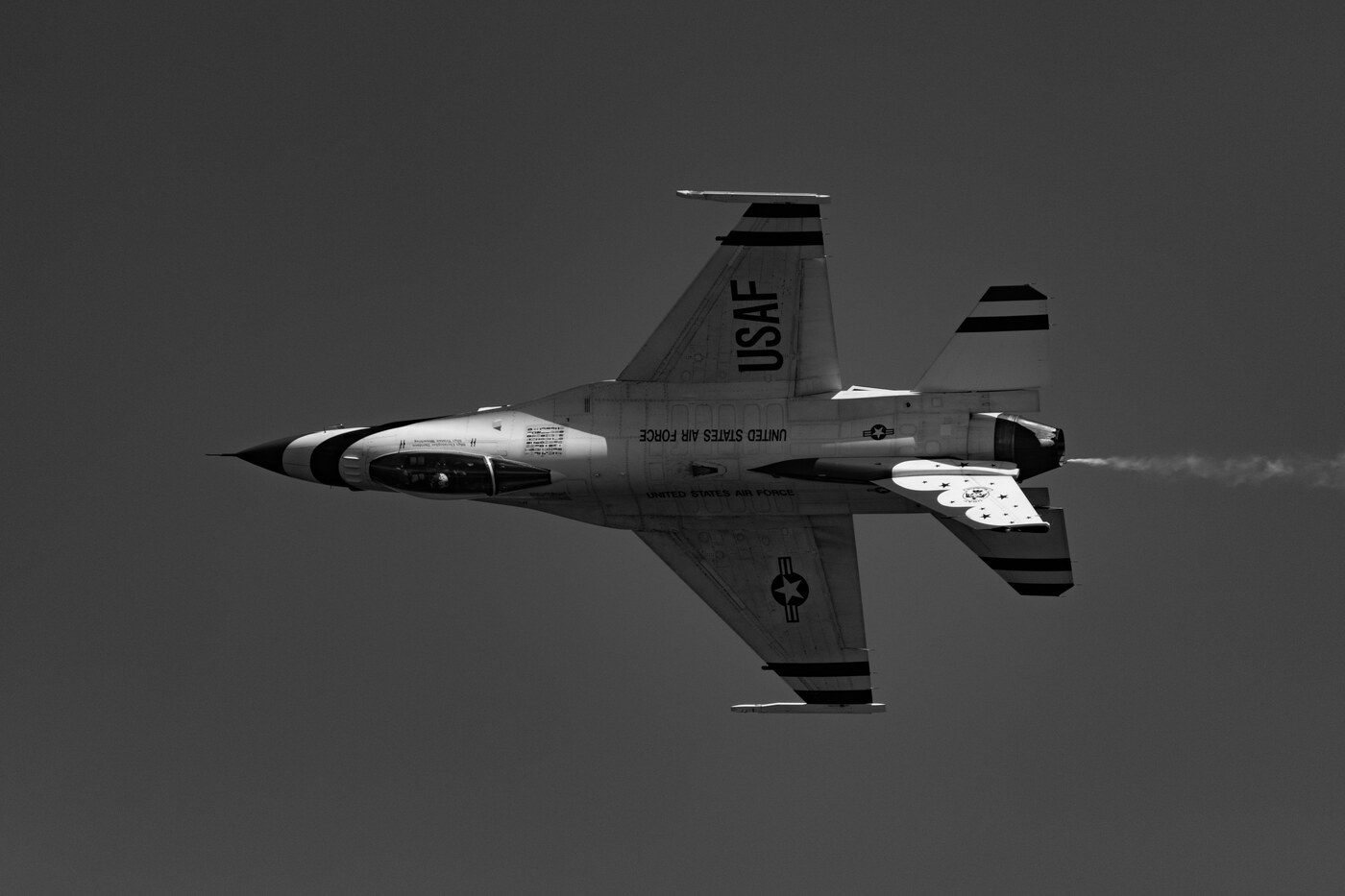 Black and white photo of a USAF Thunderbirds F-16 seen from below in a banked pass against a plain gray sky