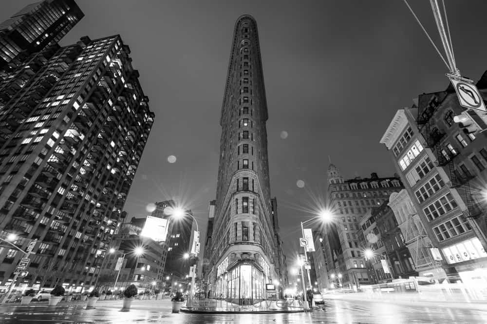 Black and white night photograph of the Flatiron Building in New York City glowing above wet streets and street lights