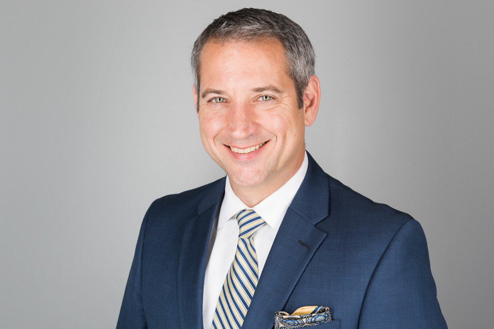Corporate headshot of a smiling man in a navy suit and striped tie against a gray background