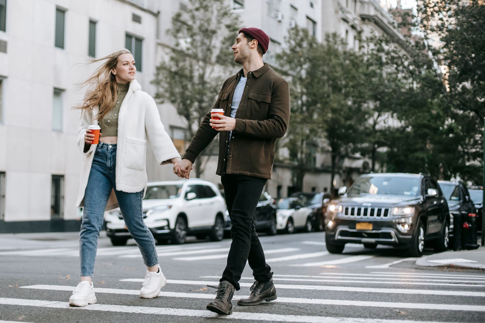 Couple holding hands and carrying coffee cups as they cross a city street
