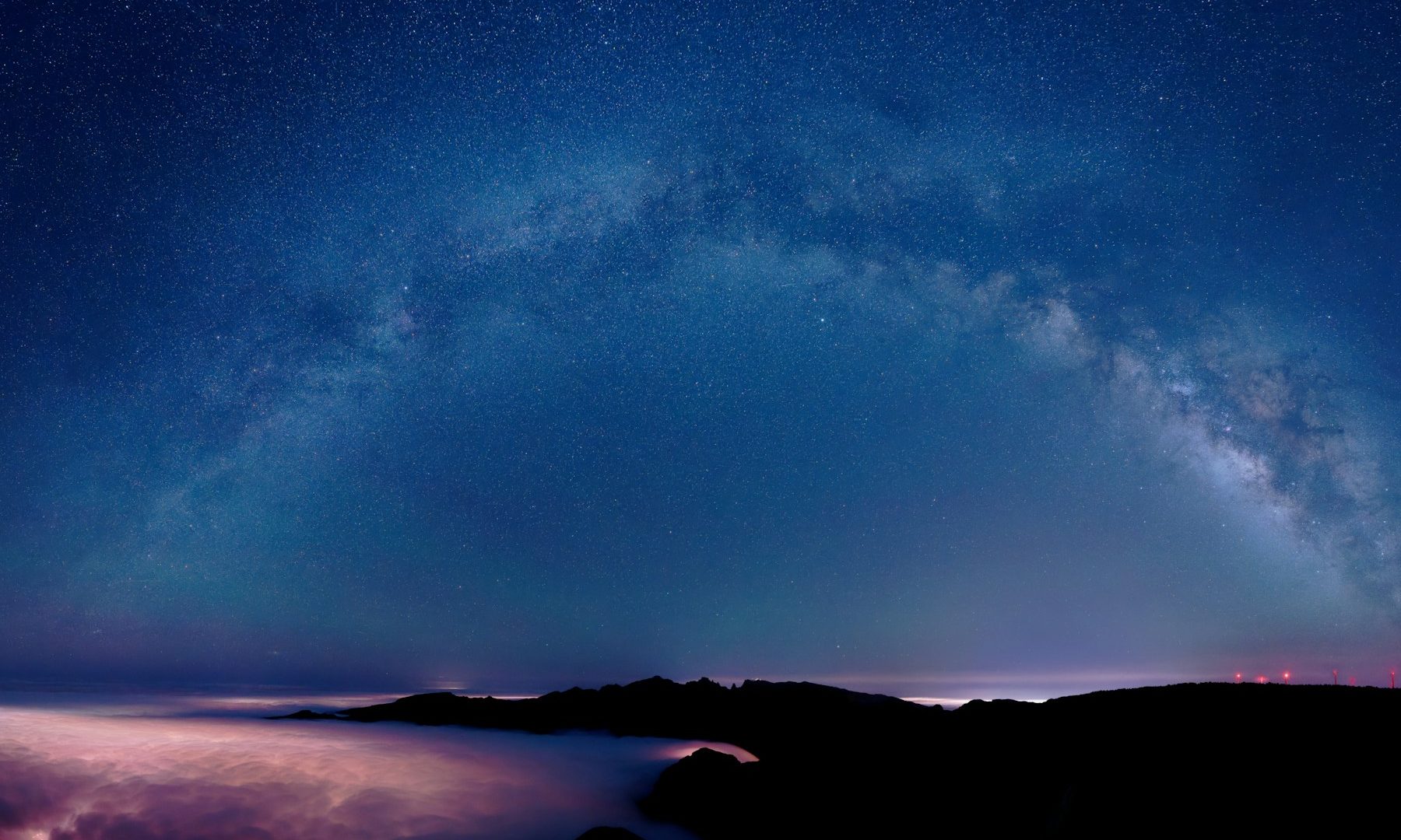 Milky Way arch and dense stars above dark mountain silhouettes and low pink clouds at night