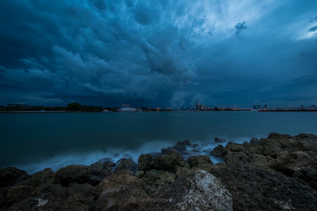 The same stormy Miami shoreline scene after editing in Lightroom, with brighter teal-blue tones, more defined storm clouds, and a glow along the horizon