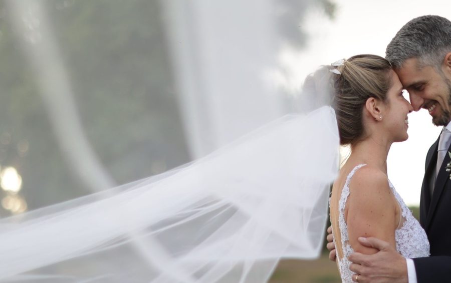 Bride and groom touching foreheads outdoors, with her veil flowing behind them.
