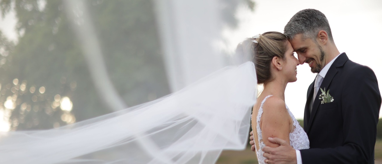 Bride and groom touching foreheads outdoors, with her veil flowing behind them.