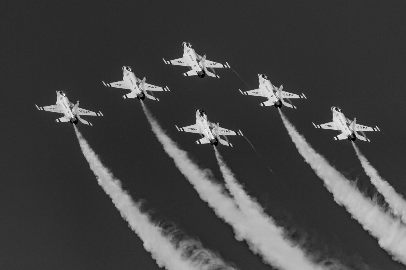Black and white photo of six USAF Thunderbirds F-16 jets spread across the frame with curving smoke trails on a dark background