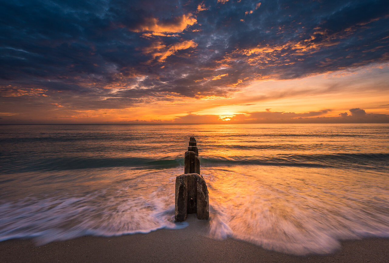 Wooden post beside breaking waves under a dramatic sunrise sky
