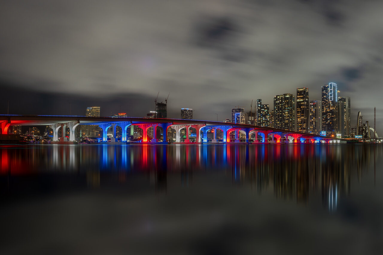 Miami bridge illuminated red and blue with city towers and reflections beneath a cloudy night sky