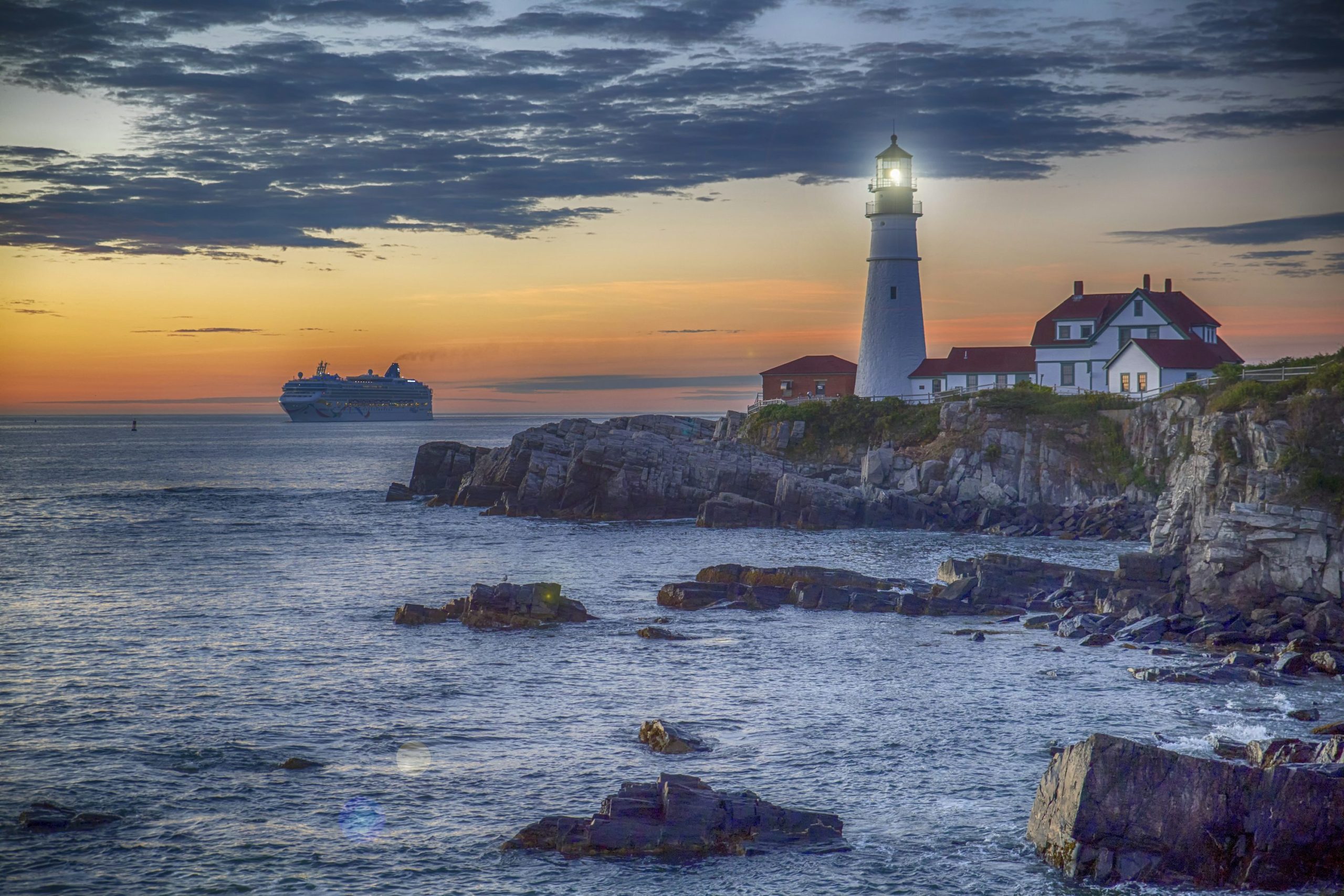 Portland Head Light and a cruise ship offshore beneath an orange twilight sky in Cape Elizabeth, Maine