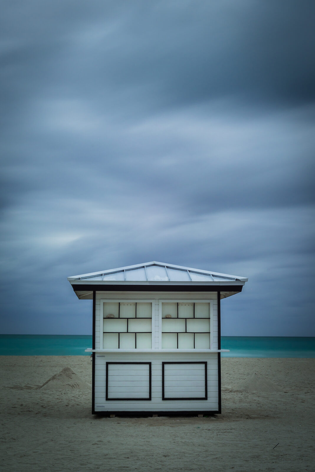 A white beach kiosk on an empty Miami beach under a dark blue-grey stormy sky and teal sea