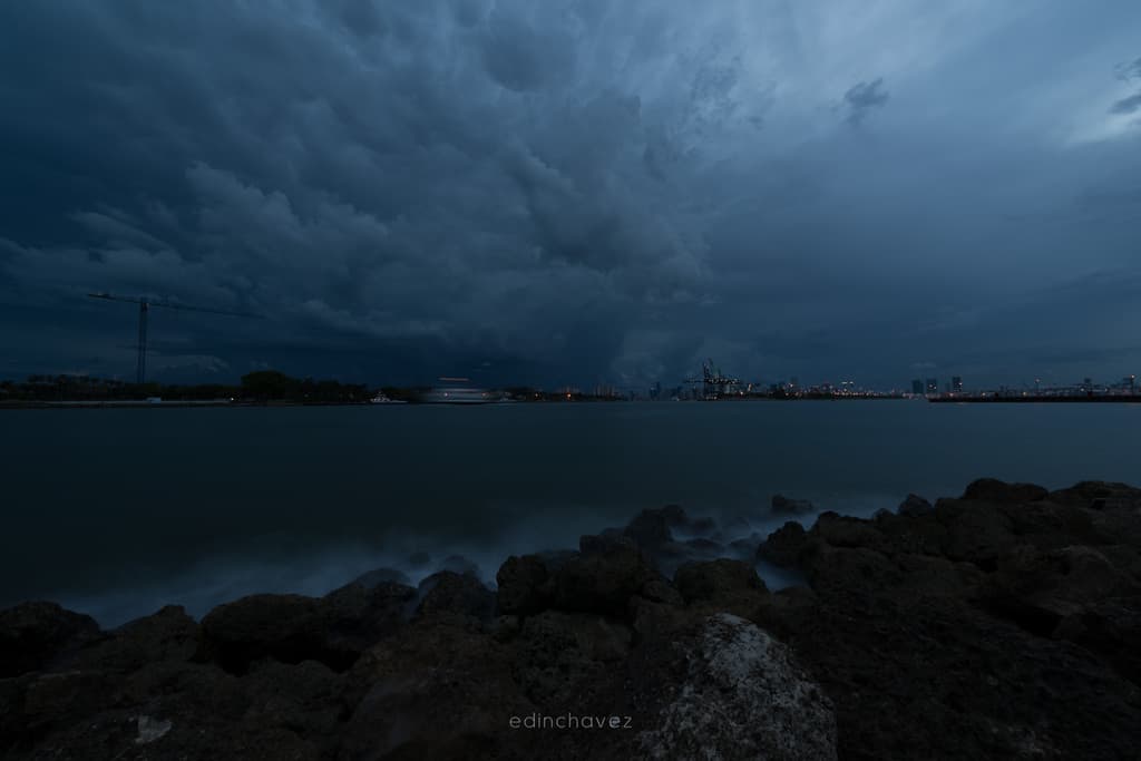 A dark, flat exposure of storm clouds over the Miami shoreline at blue hour, with rocks in the foreground and port cranes on the horizon