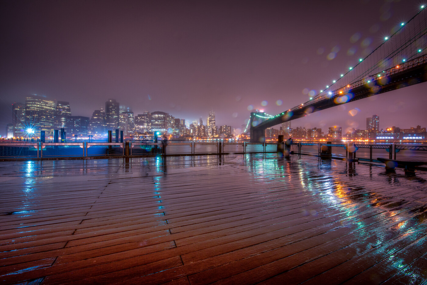 A wet wooden boardwalk at night reflecting colorful city lights with a lit bridge in the distance