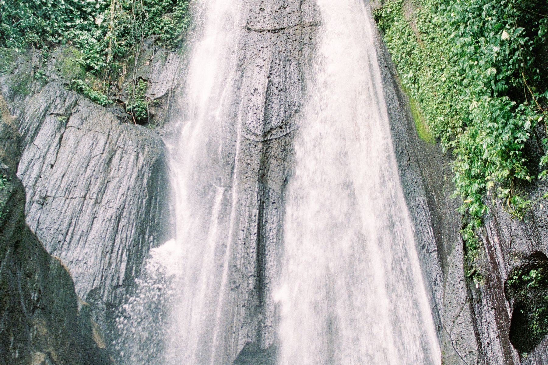 A tall waterfall running down grey rock between green jungle in Bali