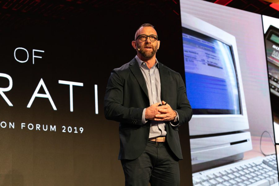 Conference speaker photographed on stage with presentation screens