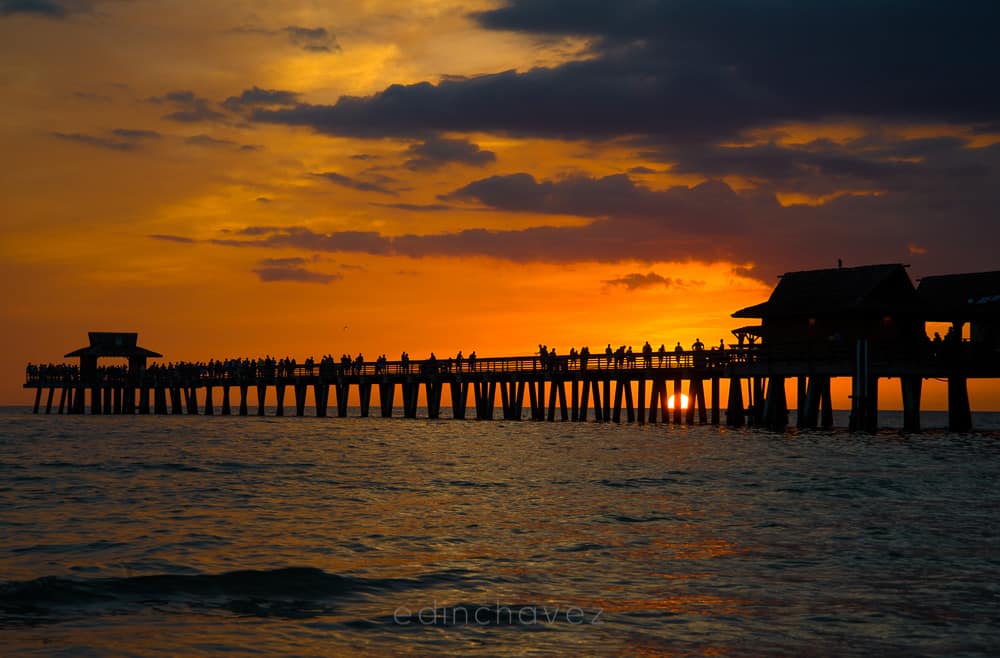 A fiery orange sunset over the Naples pier in Florida, with the sun dropping below the horizon under the pier and the crowd on it in silhouette