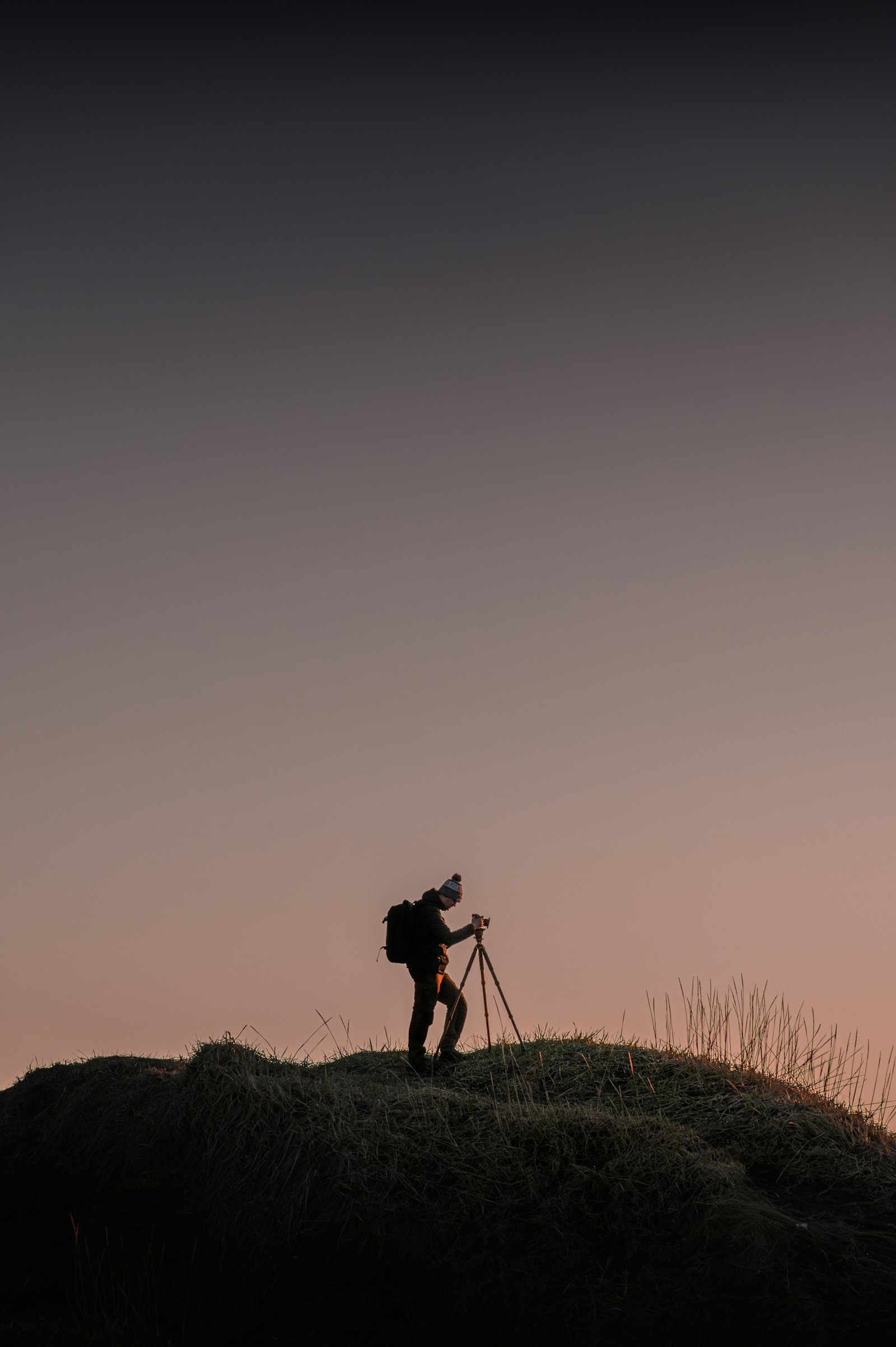 Photographer silhouetted on a grassy hilltop at dusk working with a camera on a tripod
