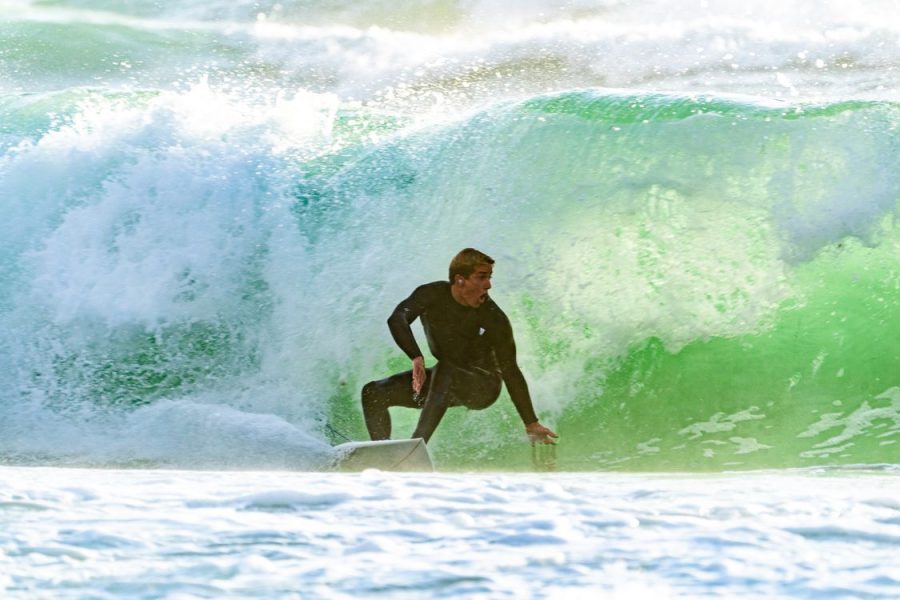 Surfer in a black wetsuit crouches on a white board inside a breaking green ocean wave.