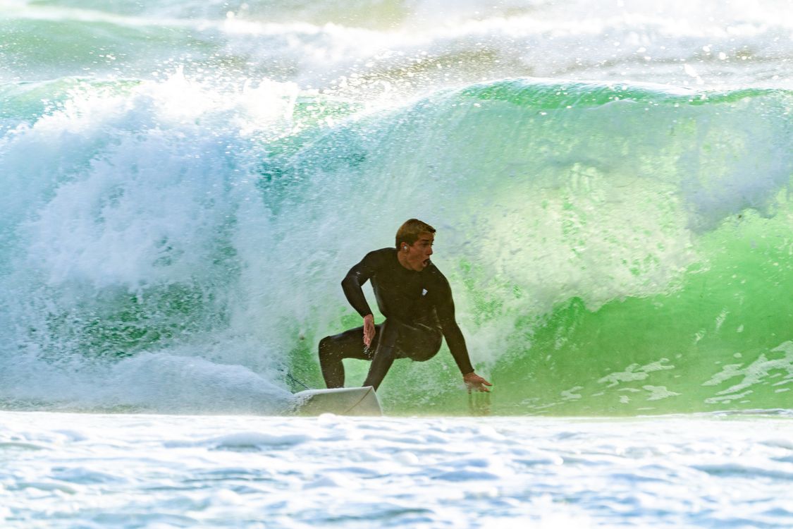 Surfer in a black wetsuit crouches on a white board inside a breaking green ocean wave.