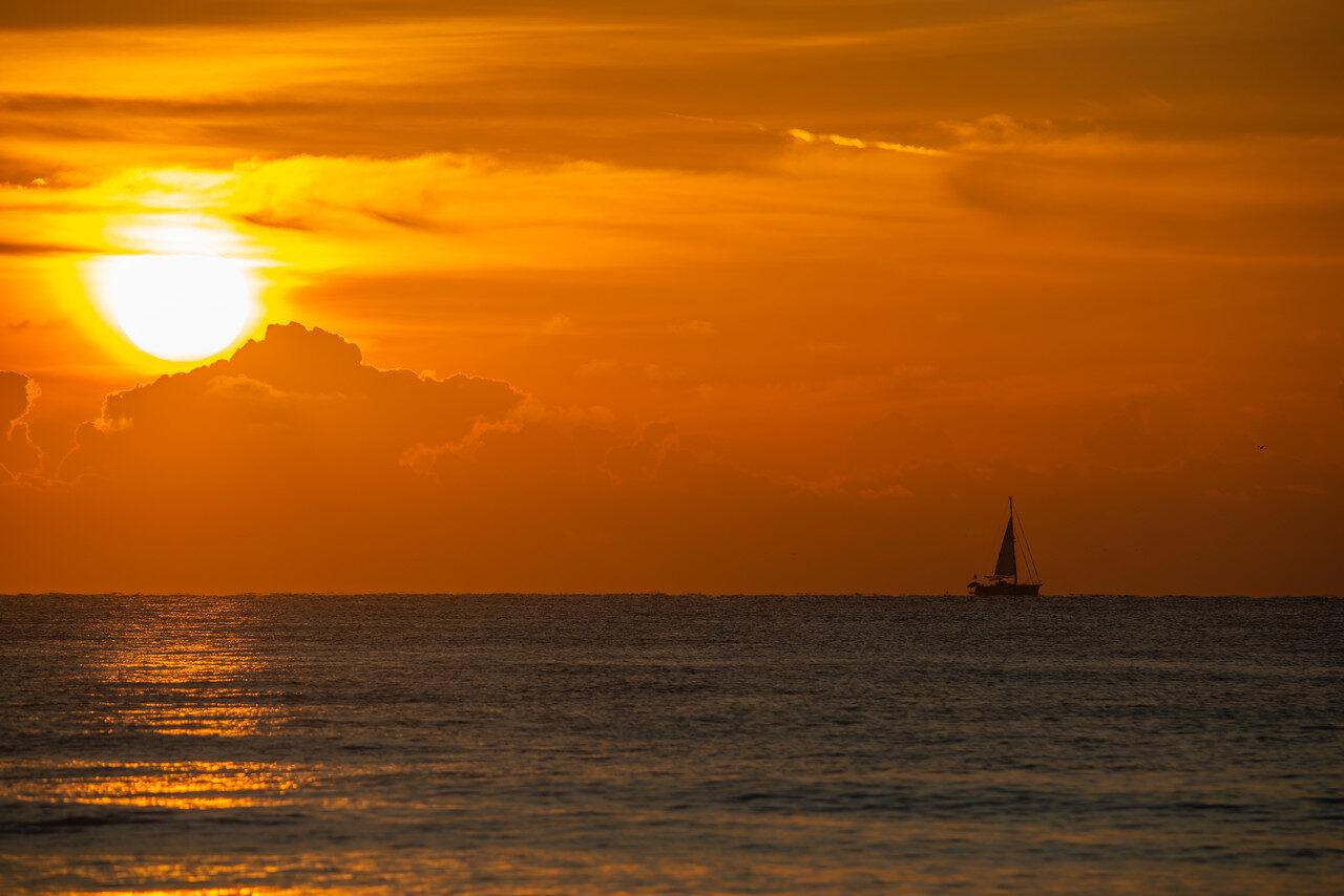 Sun disk low over a calm sea in an orange sky with a single sailboat on the horizon