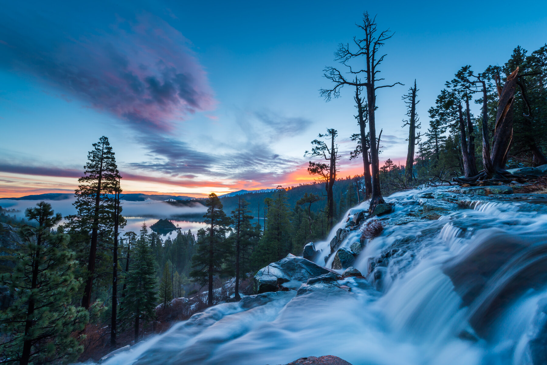 Long-exposure view of a stream of silky white water flowing over rocks below pine trees and a blue-purple dusk sky