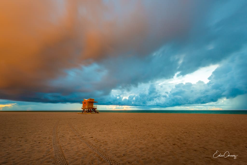 A Miami Beach lifeguard tower on empty sand under a huge sky split between warm orange storm light on the left and cool blue clouds on the right