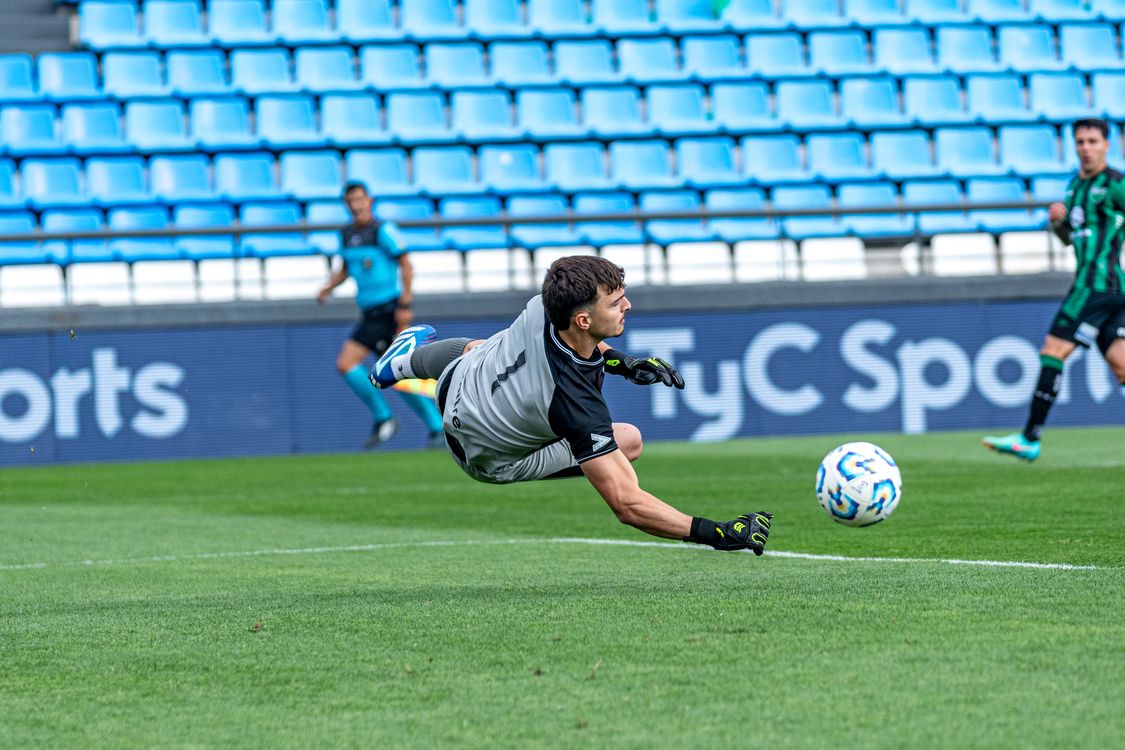 Soccer goalkeeper airborne with one glove reaching toward the ball above a green pitch.