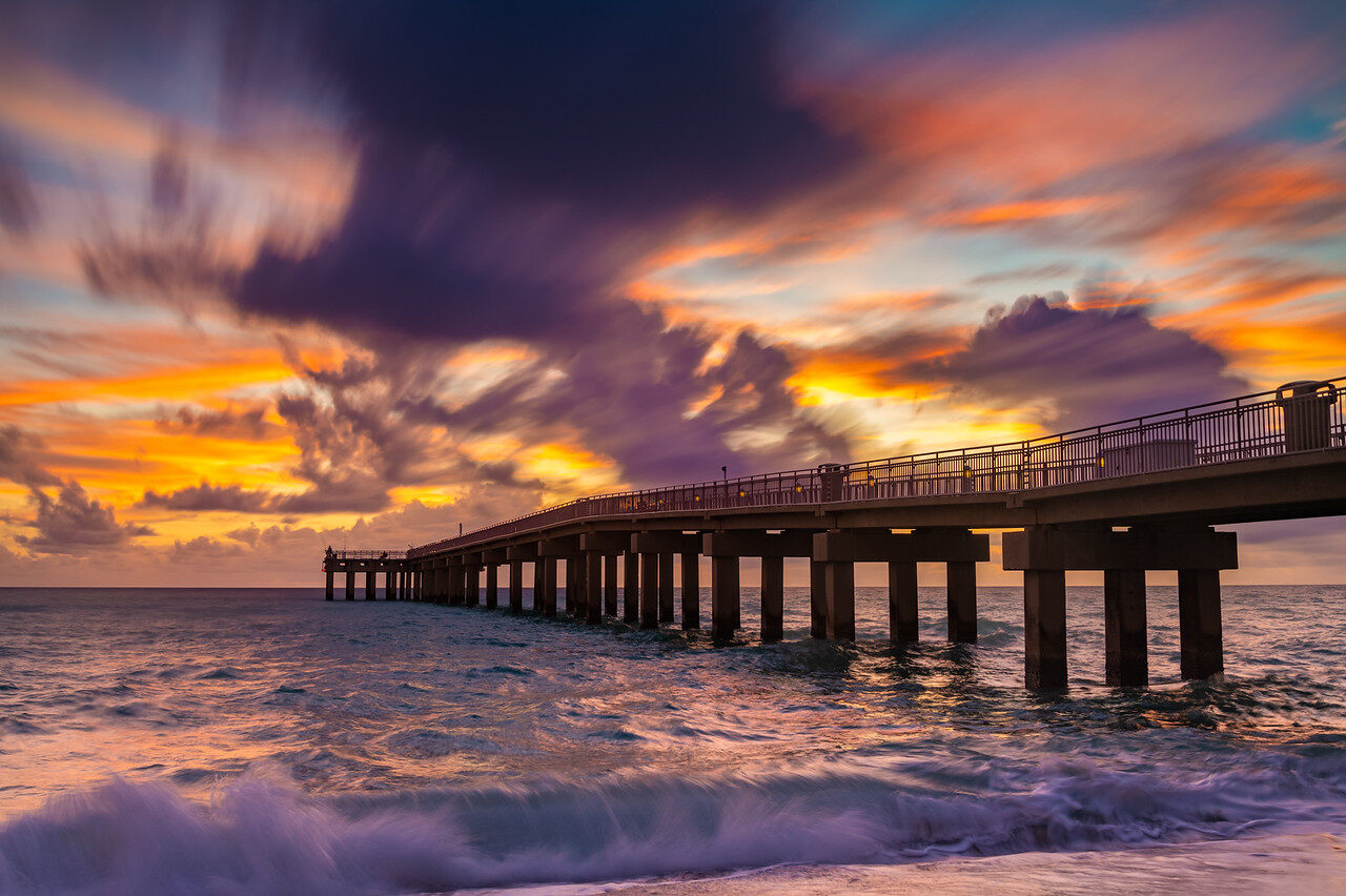 Wooden pier reaching into the sea under streaked purple and orange clouds with waves in the foreground