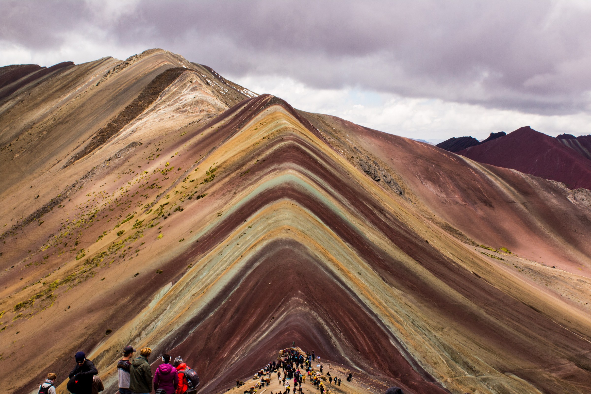 The colored slopes of Vinicunca with mountain ridges and visitors for scale.