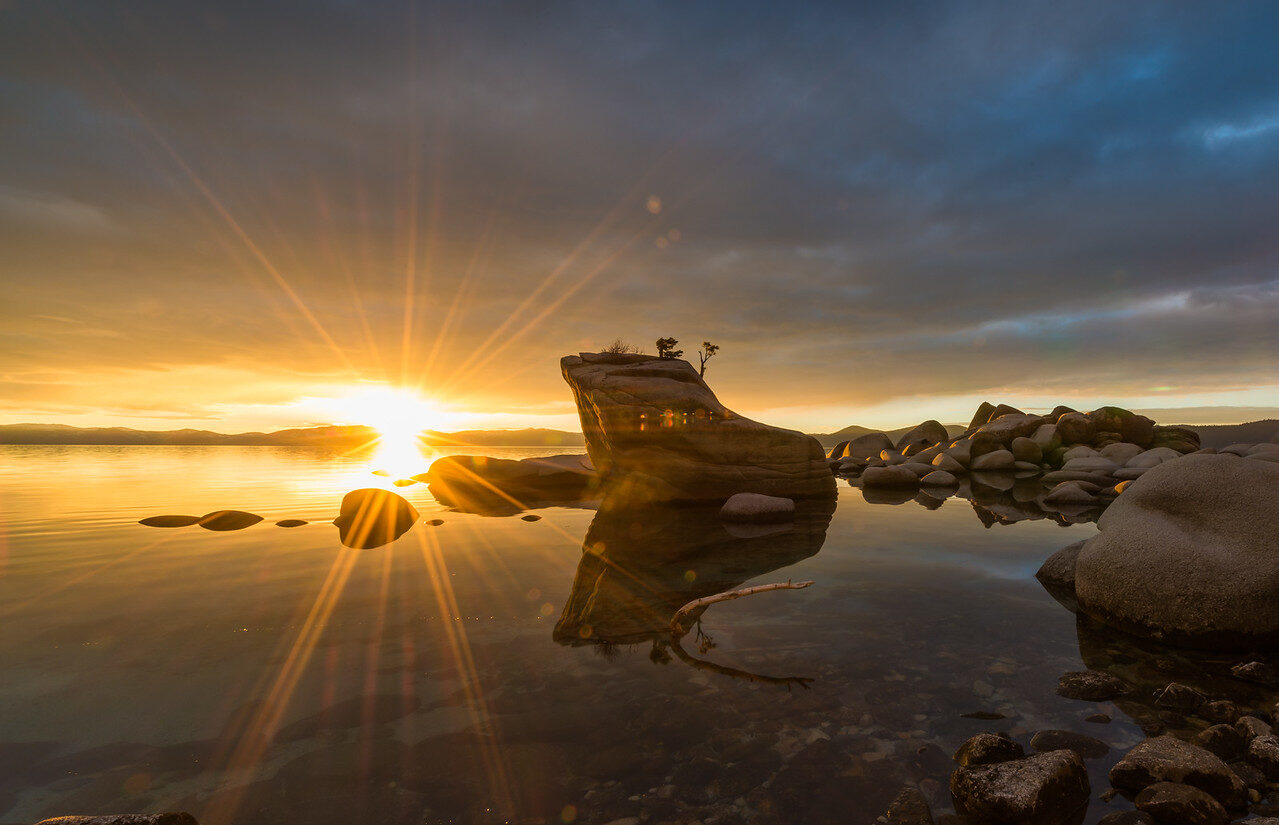 Low sun breaking into a star of rays over still water and boulders at Bonsai Rock, Lake Tahoe