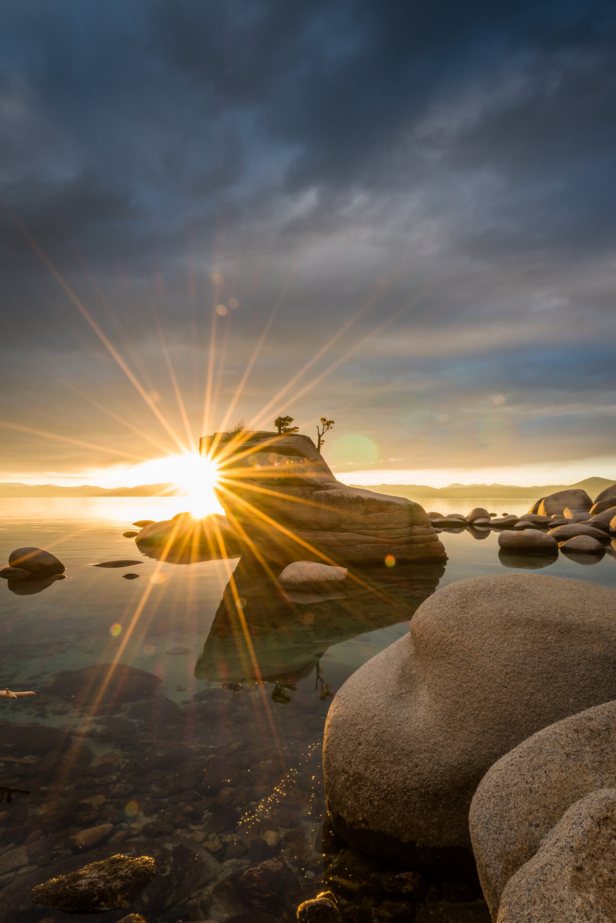 Low sun shining as a starburst behind a rock with a small tree, with large boulders in the foreground and dark clouds above