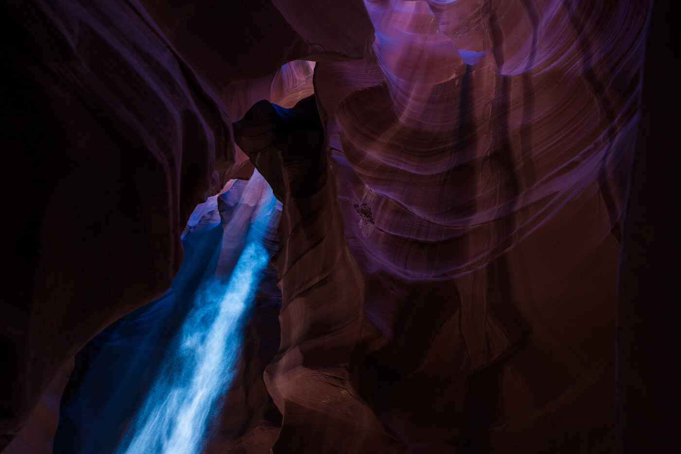 A narrow beam of light falling through a canyon opening onto curved purple and orange rock walls