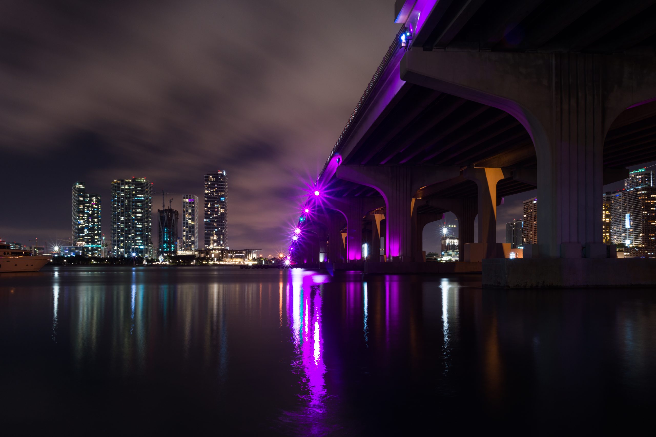 Miami skyline and purple-lit bridge over calm water at night, city lights reflected