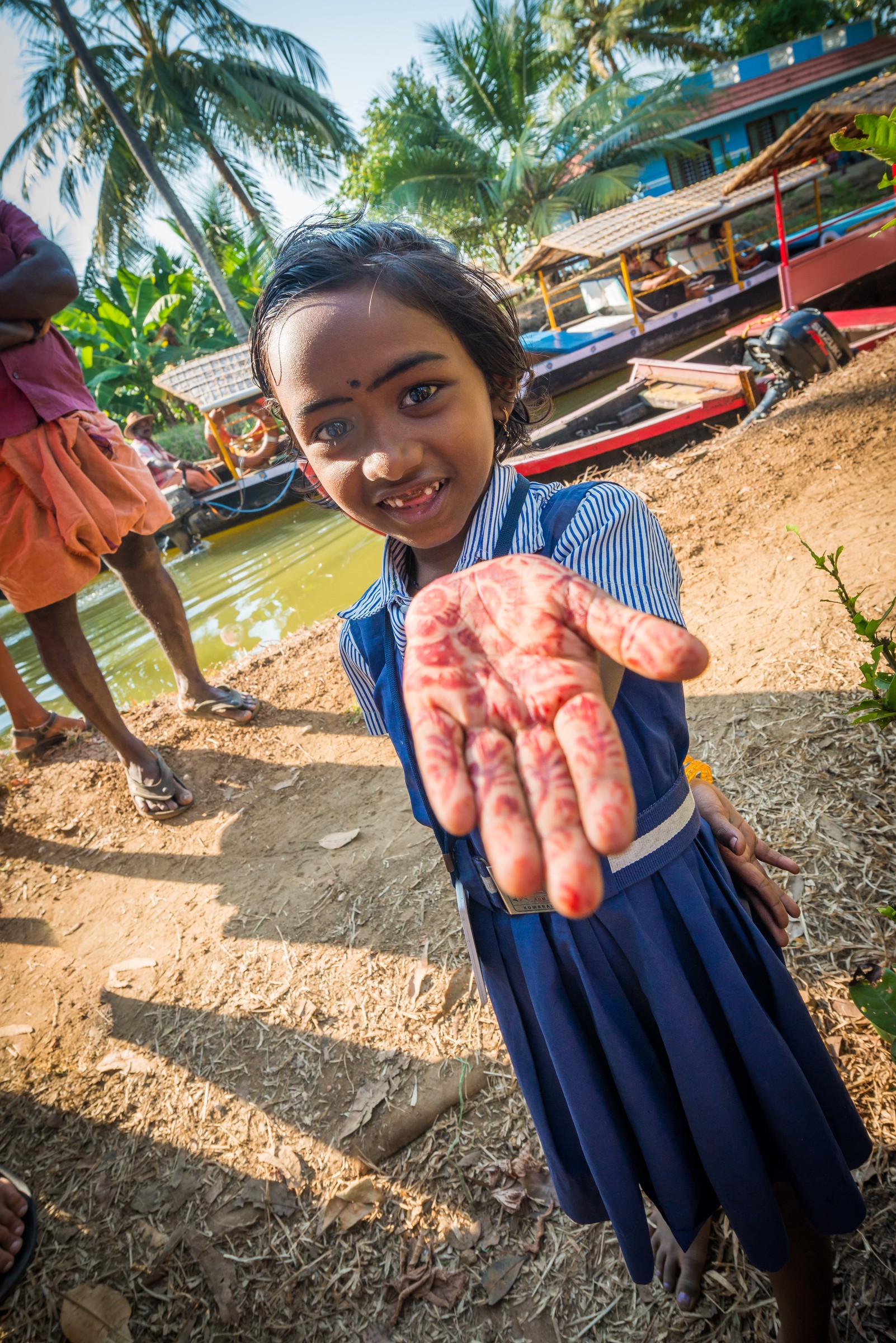 A young girl with a henna-painted hand reaching toward the camera on a Kerala backwater, her face sharp while boats and palms fall into soft blur - wide-aperture depth of field
