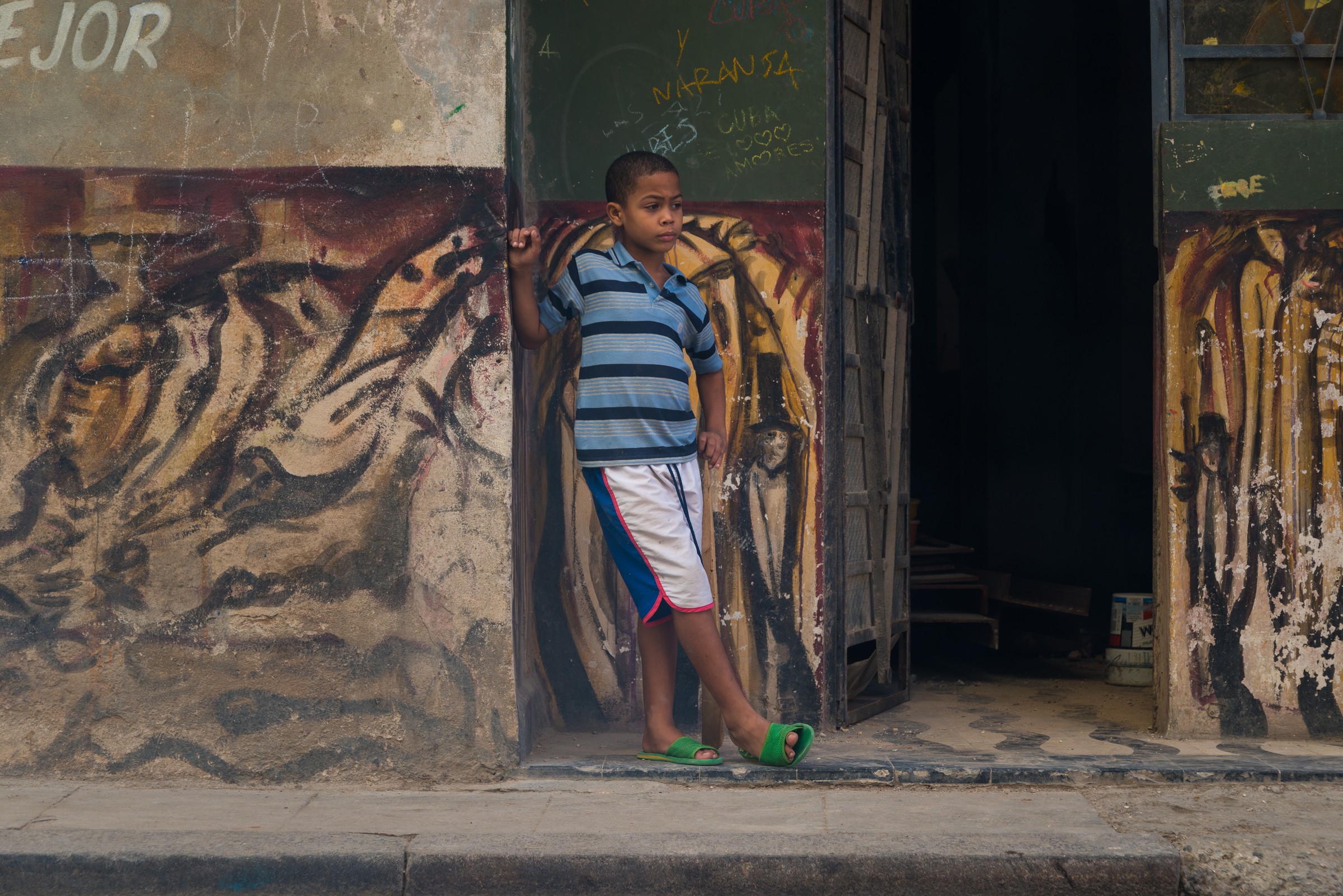 A boy standing in a shadowed Havana doorway beside a painted wall, the kind of dim scene where raising ISO buys a usable shutter speed