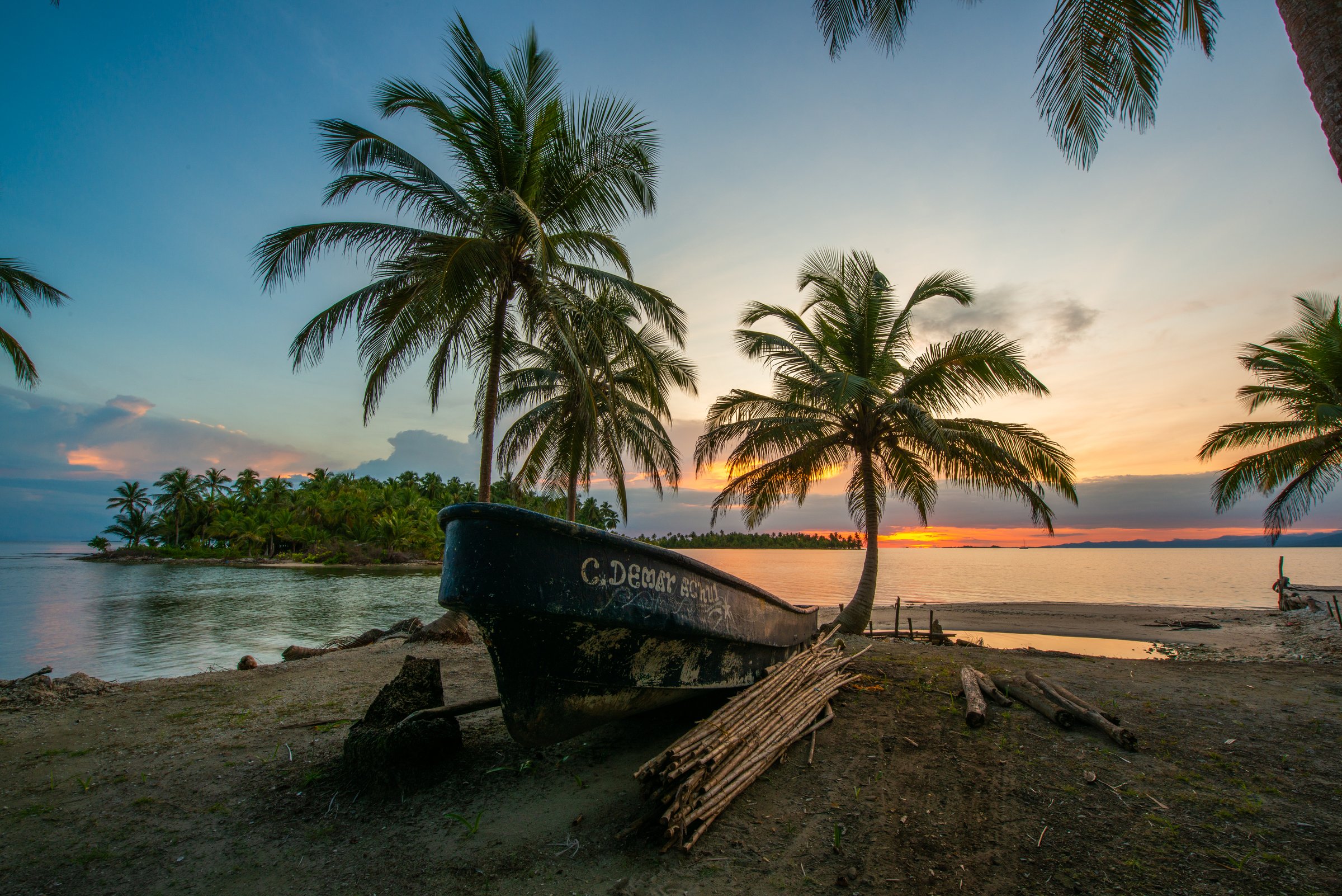 Weathered wooden boat on a sandy island shore with palm trees and a small tree-covered island at sunset